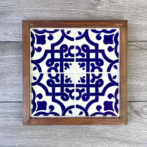 May include: Square wooden framed tile coaster. The coaster features a white background with a repeating blue geometric pattern. The frame is made of dark brown wood. The coaster is a decorative item.