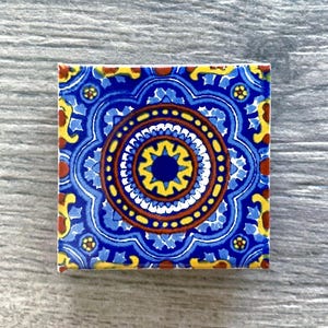 May include: A blue and yellow ceramic tile with a circular pattern in the center. The pattern features concentric circles of red, white, and yellow, with a blue star in the center. The tile has a decorative border with a floral design.