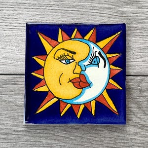 May include: Square ceramic tile with a celestial design. The tile features a yellow sun and a white moon with blue eyes and red lips, set against a dark blue background. The sun has orange and yellow rays.