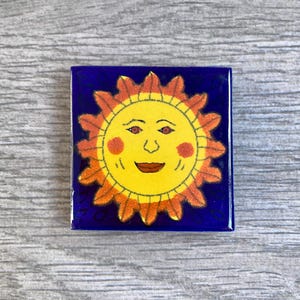 May include: A ceramic tile with a blue background and a yellow sun with a smiling face. The sun has red cheeks and a red mouth. The sun has orange rays.