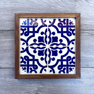 May include: Square decorative tile with a blue and white floral pattern, set in a dark brown wooden frame. The tile design features a symmetrical arrangement of stylized flowers and geometric shapes, creating a classic, elegant look.