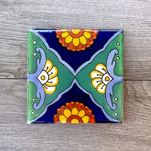 Pode incluir: Um azulejo de cerâmica quadrado com um padrão floral colorido. O azulejo apresenta um fundo azul com desenhos florais verdes, amarelos, laranja e brancos.