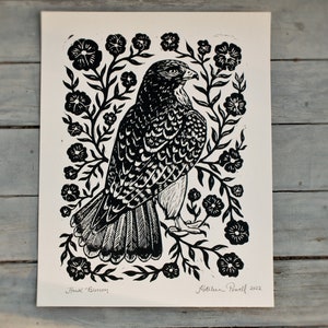 May include: Black and white linocut print of a hawk surrounded by floral elements. The artwork is titled "Hawk Blossom" and signed by the artist, Kathleen Powell, with the year 2022.