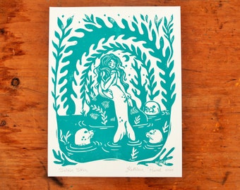 May include: A teal blue linocut print of a mermaid with long hair and a tail, surrounded by seaweed and three seals. The print is titled "Selkie Skin" and signed "Yilleen Powell 2020".