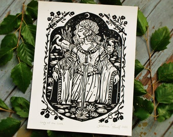May include: Black and white woodblock print of a woman standing in a lake holding a sword. She is surrounded by water lilies and reeds. The print is titled "Lady of the Lake" and signed "Kathleen Powell 2022".
