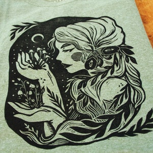 May include: Black and white illustration of a woman with long hair holding a cluster of mushrooms in her hand. The woman is surrounded by leaves and vines. The illustration is in a woodcut style.