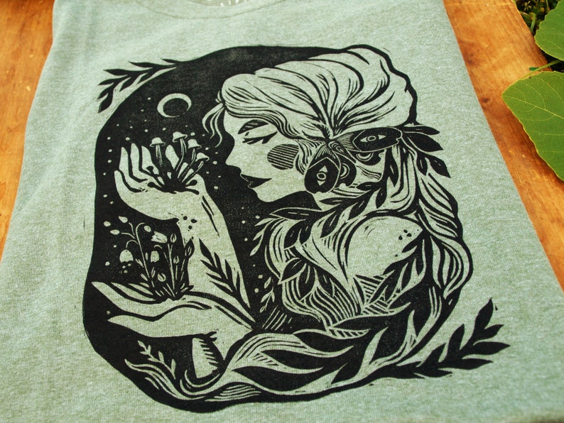 May include: Black and white illustration of a woman with long hair holding a cluster of mushrooms in her hand. The woman is surrounded by leaves and vines. The illustration is in a woodcut style.