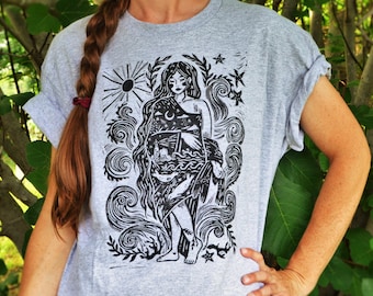 May include: A light gray t-shirt featuring a black and white illustration of a woman with long hair, a sun, moon, stars, and ocean waves. The design includes a lighthouse and other nautical elements. The sleeves are rolled up.