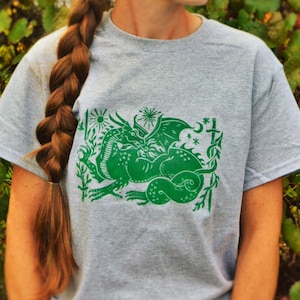 Puede incluir: Una camiseta gris jaspeada con un estampado verde de un dragón y una serpiente entrelazados. El estampado incluye un sol, una luna, estrellas y una espada.