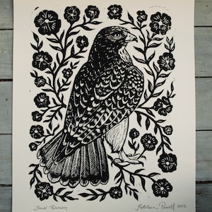 May include: Black and white linocut print of a hawk surrounded by floral elements. The artwork, titled "Hawk Blossom," features intricate details and is signed by the artist, Kathleen Powell, with the year 2022.