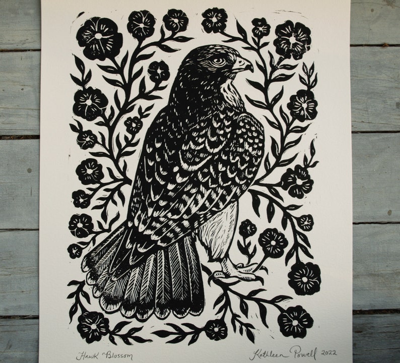 May include: Black and white linocut print of a hawk surrounded by floral elements. The artwork, titled "Hawk Blossom," features intricate details and is signed by the artist, Kathleen Powell, with the year 2022.