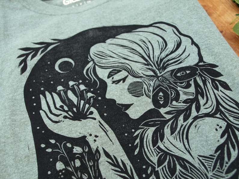 May include: Black and white illustration of a woman with long hair, holding a cluster of mushrooms in her hand. The woman is surrounded by leaves and flowers. The illustration is on a light green background.