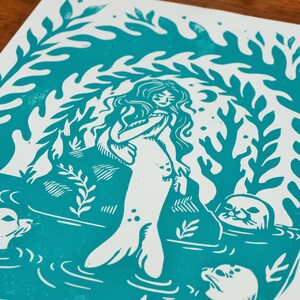 May include: A teal blue linocut print of a mermaid with long hair and a tail, surrounded by seaweed and three seals. The mermaid is standing on a rock and looking to the right.