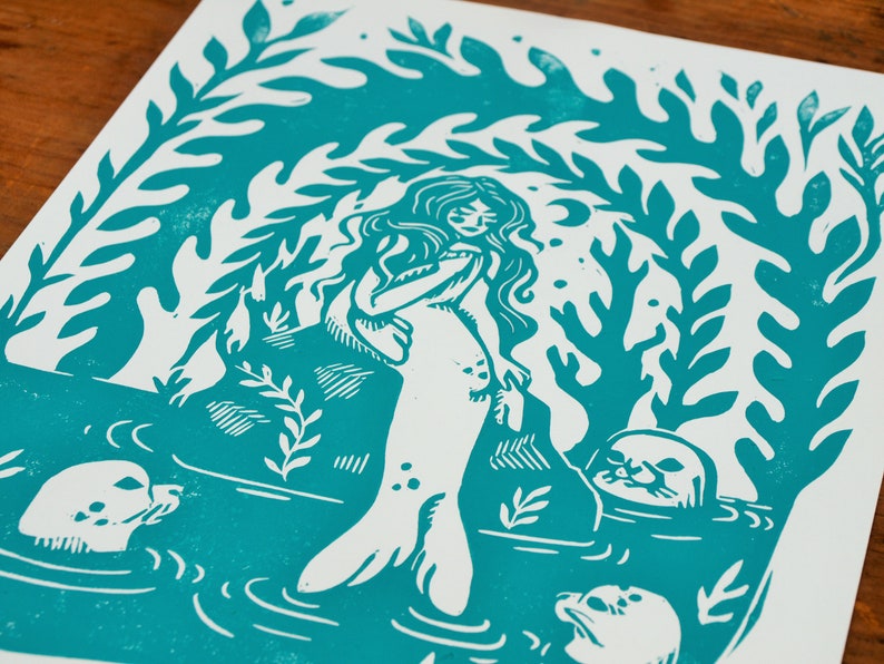 May include: A teal blue linocut print of a mermaid with long hair and a tail, surrounded by seaweed and three seals. The mermaid is standing on a rock and looking to the right.