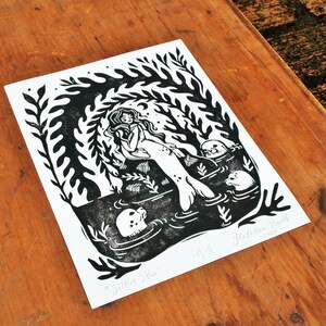 May include: Black and white linocut print of a mermaid with three seals. The mermaid is sitting on a rock with her tail in the water. The print is titled "Selkie Ska" and signed "Kathleen Powell 2015".