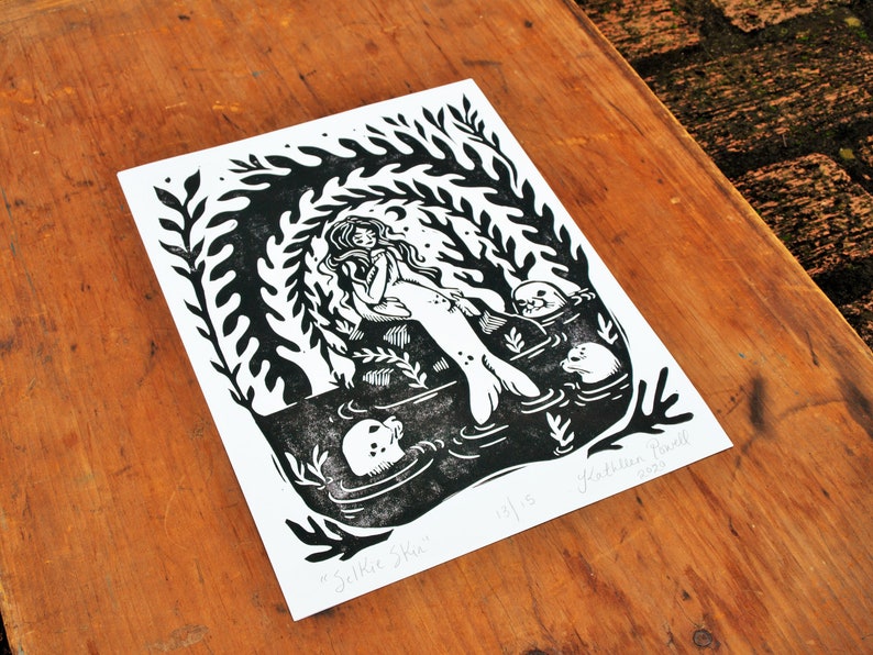 May include: Black and white linocut print of a mermaid with three seals. The mermaid is sitting on a rock with her tail in the water. The print is titled "Selkie Ska" and signed "Kathleen Powell 2015".
