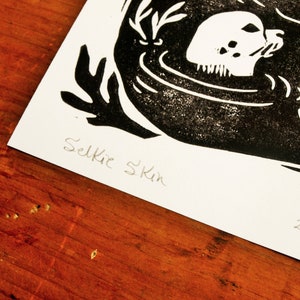 May include: Black and white linocut print of a seal with the text "Selkie Skin" and the number "2/5" in the bottom right corner.
