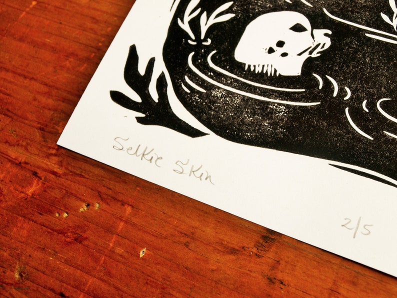 May include: Black and white linocut print of a seal with the text "Selkie Skin" and the number "2/5" in the bottom right corner.