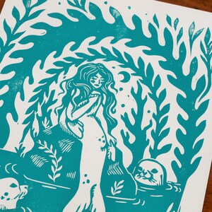 May include: A teal blue linocut print of a mermaid with long hair and a seal. The mermaid is surrounded by seaweed and coral.