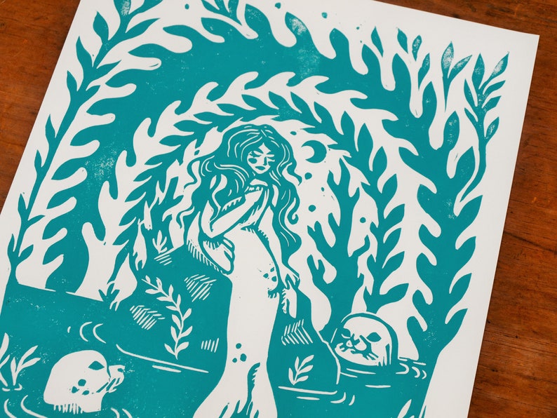 May include: A teal blue linocut print of a mermaid with long hair and a seal. The mermaid is surrounded by seaweed and coral.