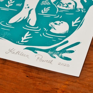 May include: Teal blue linocut print of a seal with its flippers in the air, surrounded by other seals and seaweed. The print is signed "Kathleen Powell 2020".