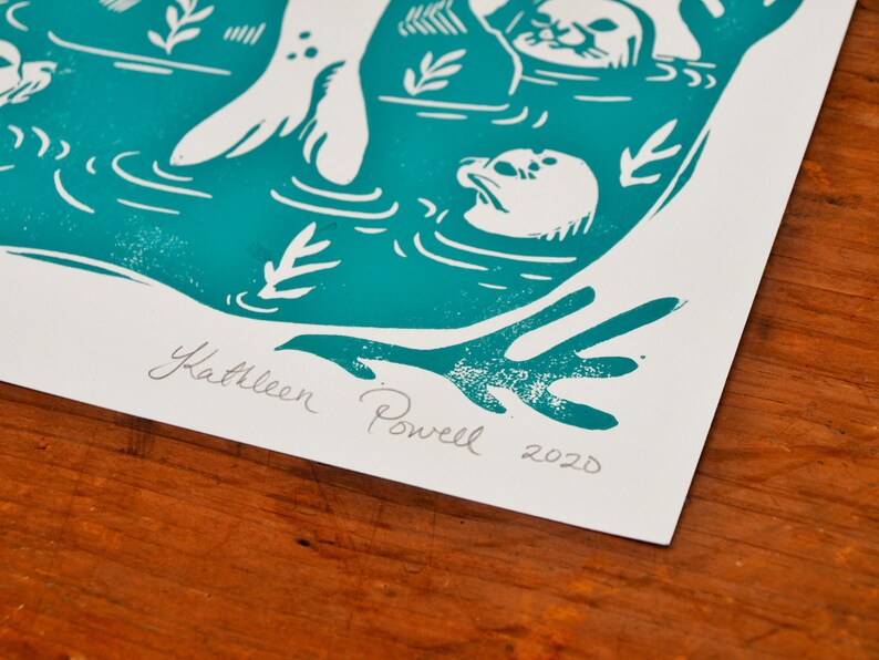 May include: Teal blue linocut print of a seal with its flippers in the air, surrounded by other seals and seaweed. The print is signed "Kathleen Powell 2020".