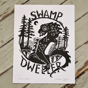 Può includere: Stampa artistica in bianco e nero con un'illustrazione di una creatura della palude. L'opera include il testo "SWAMP DWELLER" in un design circolare, con alberi e una luna. Firmata dall'artista, Kathleen Powell, 2023.