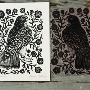 May include: Black and white linocut print of a hawk surrounded by floral elements. The print is on white paper, with the linocut block next to it. The hawk is detailed with textured feathers. The print is signed "Hawk Blossom" and "Kathleen Powell 2022".