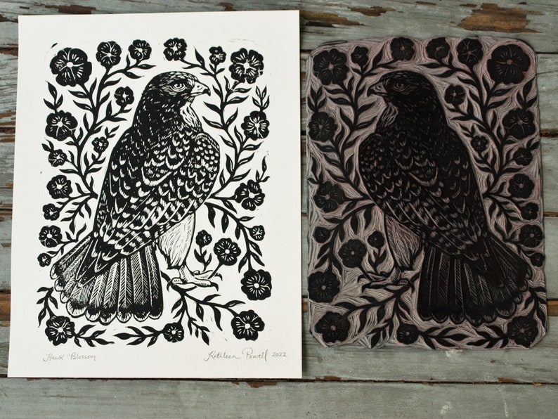 May include: Black and white linocut print of a hawk surrounded by floral elements. The print is on white paper, with the linocut block next to it. The hawk is detailed with textured feathers. The print is signed "Hawk Blossom" and "Kathleen Powell 2022".
