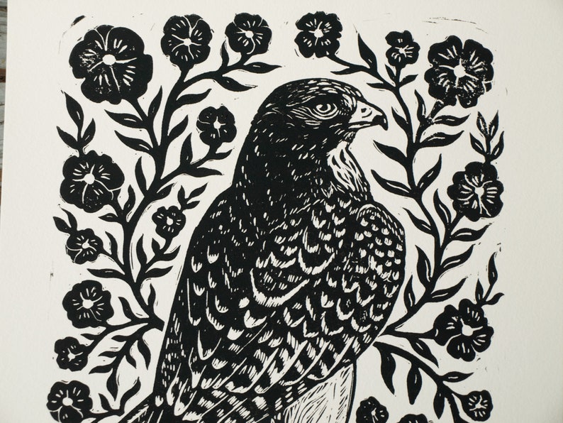 May include: Black and white linocut print of a falcon surrounded by floral elements. The bird is depicted in profile, with detailed feather patterns. The flowers and foliage create a decorative border around the bird, giving the print a natural theme.