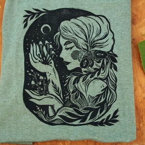 May include: A black and white illustration of a woman with a floral crown and a moth on her shoulder. She is holding a cluster of mushrooms in her hand. The illustration is on a green background.