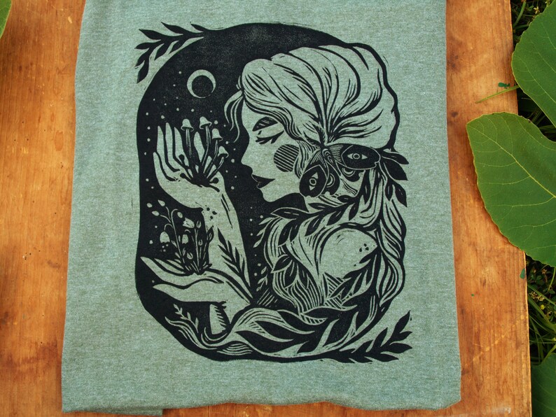 May include: A black and white illustration of a woman with a floral crown and a moth on her shoulder. She is holding a cluster of mushrooms in her hand. The illustration is on a green background.