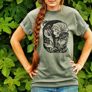 May include: A green t-shirt with a black and white illustration of a woman with flowers and a crescent moon. The woman is surrounded by leaves and vines.