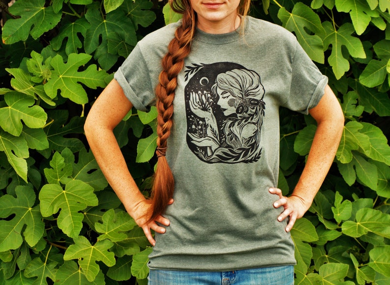 May include: A green t-shirt with a black and white illustration of a woman with flowers and a crescent moon. The woman is surrounded by leaves and vines.