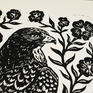 May include: Black and white linocut print of a hawk with detailed feathers and a sharp beak, surrounded by stylized floral elements. The artwork is a close-up, showcasing the bird's profile and the texture of the print.