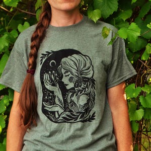 May include: A green t-shirt with a black and white illustration of a woman with long hair and a floral crown. The woman is holding a bouquet of flowers and is surrounded by leaves and vines.