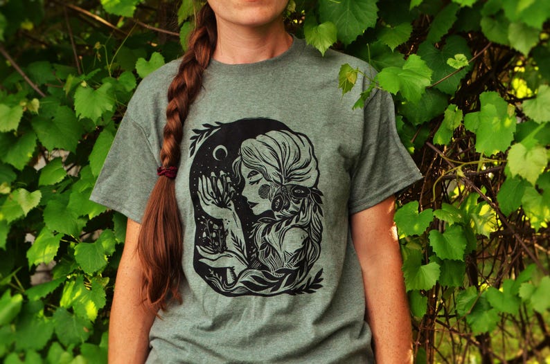 May include: A green t-shirt with a black and white illustration of a woman with long hair and a floral crown. The woman is holding a bouquet of flowers and is surrounded by leaves and vines.