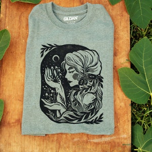 May include: A heather grey t-shirt with a black and white illustration of a woman with long hair surrounded by foliage and holding a cluster of mushrooms. The text "Gildan" is visible on the shirt.