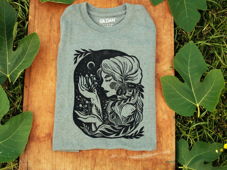 May include: A heather grey t-shirt with a black and white illustration of a woman with long hair surrounded by foliage and holding a cluster of mushrooms. The text "Gildan" is visible on the shirt.