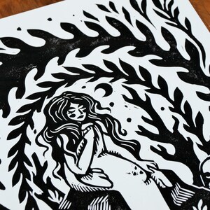 May include: Black and white linocut print of a woman with long hair sitting under a leafy canopy. The woman is wearing a simple dress and has her eyes closed. A small animal is sitting beside her.