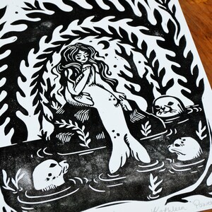 May include: Black and white linocut print of a mermaid sitting on a rock with three seals swimming around her. The print is signed "Kathleen Powell 2020".