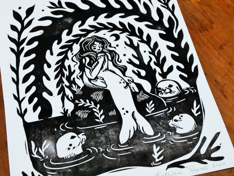 May include: Black and white linocut print of a mermaid sitting on a rock with three seals swimming around her. The print is signed "Kathleen Powell 2020".