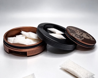 May include: A round, metallic container with a copper-coloured base and a black lid featuring an engraved design. The container is open, revealing several small, white, rectangular pouches inside, with two additional pouches resting outside.