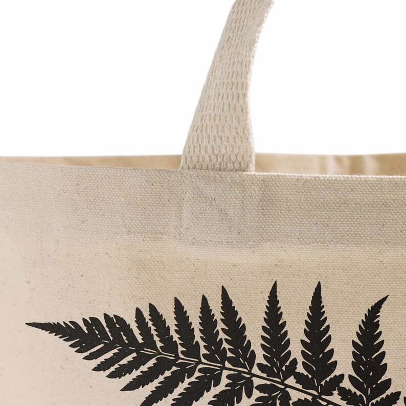Material close-up on another natural canvas tote showing a woven handle, top seam, and black fern print.