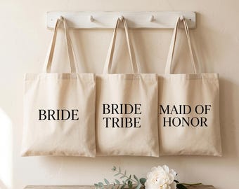 Puede incluir: Tres bolsas de mano color crema con texto negro que dice "BRIDE", "BRIDE TRIBE" y "MAID OF HONOR". Las bolsas cuelgan de un estante blanco en una pared. Un banco de madera sostiene una peonía blanca y vegetación.
