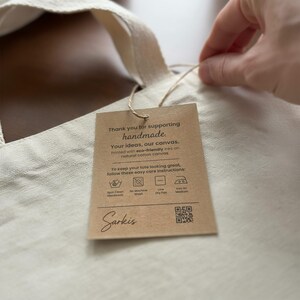 Hand attaching a Bijouland care tag to a natural cotton canvas tote; the visible label includes eco-friendly ink wording and a QR code.