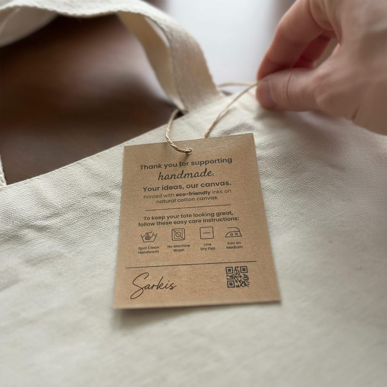 Hand attaching a Bijouland care tag to a natural cotton canvas tote; the visible label includes eco-friendly ink wording and a QR code.