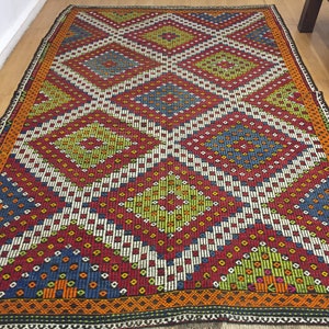 May include: A colorful rug with a geometric pattern of diamonds and squares in shades of red, blue, green, and white. The rug has a fringe border.