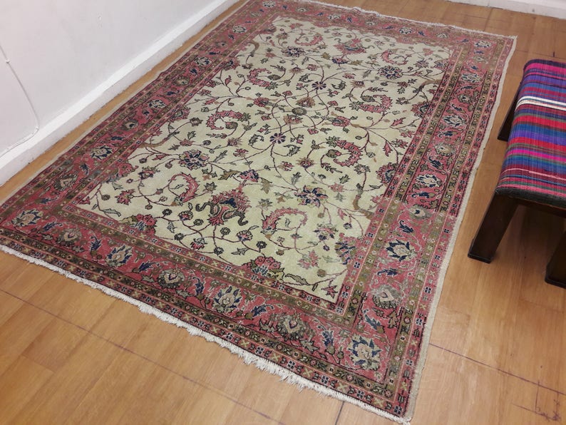 May include: A cream-colored rug with a floral pattern in shades of pink, green, and blue. The rug has a fringed edge and a border with a repeating pattern.