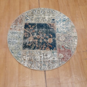May include: A round, multi-colored rug with a patchwork design. The rug has a cream background with blue, red, and green accents. The rug is made of woven fabric and has a fringed edge.